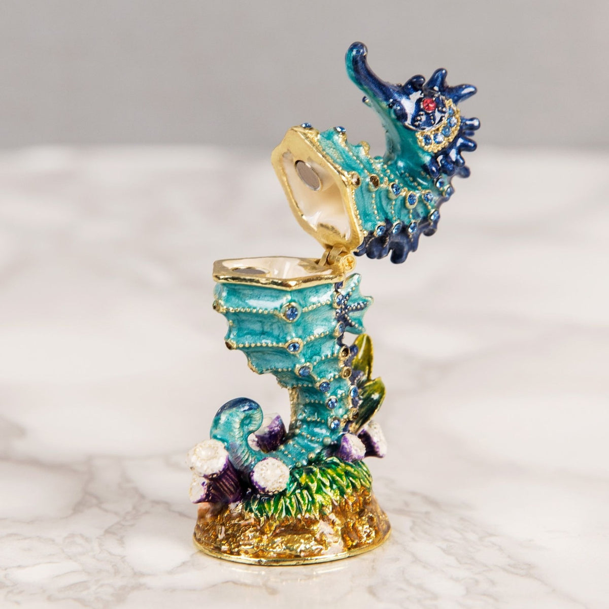 Sea Horse hand painted metal trinket box with hinged lid, turquoise and navy blue body with gold detailing and blue stones, on a gold base with coral, shown open against a light background.
