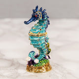 Sea Horse Hand Painted Metal Trinket Box with hinged lid, turquoise ridged body, navy blue crested head, jewelled accents on a gold-toned seaweed base, set against a marbled background.