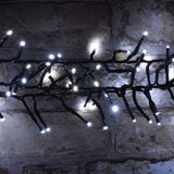 LYYT 960 LED outdoor string lights with cool white bulbs clustered along a dark green cable, draped against a rustic whitewashed brick wall.