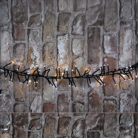 The LYYT 960 LED Outdoor String Lights - Dual Colour, featuring both white and yellow bulbs, are elegantly draped across a textured stone wall, creating a warm and cozy ambiance. With an IP44 rating, they are designed for durability in both indoor and outdoor settings.