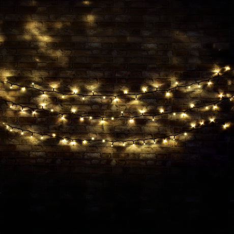 The LYYT 90 LED Heavy Duty LED String Lights, 10m - Warm White, are elegantly draped in gentle arcs against a dark brick wall, crafting a cozy and inviting atmosphere. These energy-efficient lights softly illuminate the textured surface, offering both charm and sustainability.