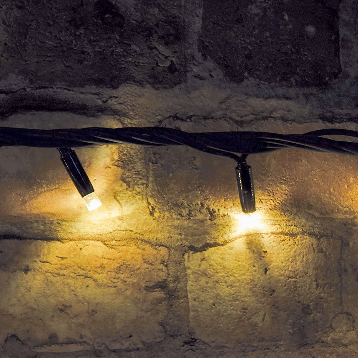 LYYT warm white LED festoon lights by AVSL draped along a rustic stone wall, showing two lit bulbs on the black rubber cable.