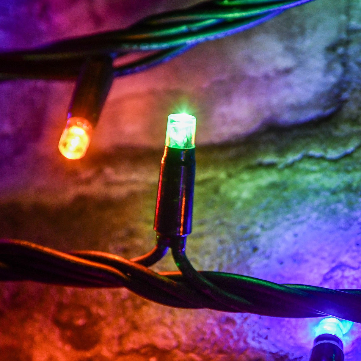 Close-up of a LYYT RGBA LED string light showing green and amber concave bulbs on heavy-duty black rubber cable against a textured stone surface.