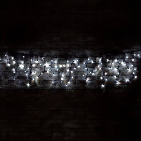 The LYYT 100 LED Outdoor Connectable Icicle String Lights, measuring 2 meters in length and emitting a cool white glow, are strung against a dark brick wall. These energy-efficient lights create a gentle, shimmering effect, as the LEDs form an intricate icicle pattern that softly illuminates the textured surface of the bricks.