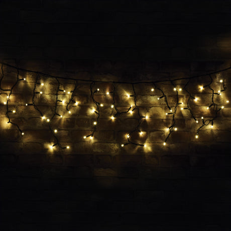 The LYYT 100 LED Outdoor Connectable Icicle String Light, 2m - Warm White, casting a warm glow against a dark brick wall, creates a cozy and festive ambiance. Designed for outdoor use, these energy-efficient lights provide the perfect illumination while maintaining low power consumption.