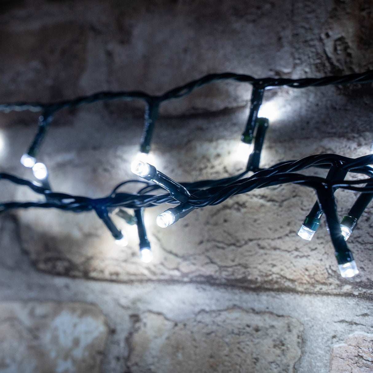 LYYT 200TS-CW cool white LED string lights on a dark green cable, draped against a rustic stone wall with bright white LEDs illuminated.