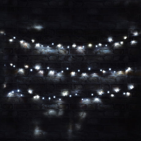 The LYYT 200 LED Indoor/Sheltered Outdoor String Lights with a cool white glow hang against a dark brick wall. Their energy-efficient design creates a cozy and festive atmosphere, with their soft illumination highlighting the rough texture of the bricks. These string lights feature a 24-hour auto-timer for added convenience and stretch over a length of 10 meters.