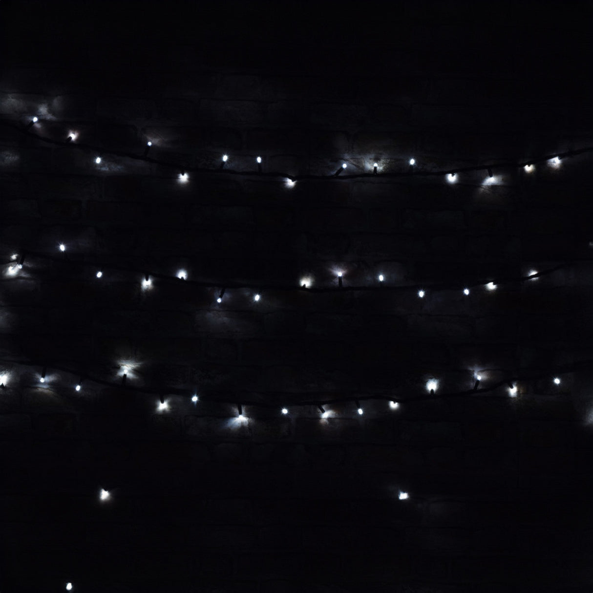 The LYYT 90 LED Heavy Duty String Lights with Controller, featuring 11 meters of cool white LEDs, are suspended in the darkness to form a pattern of glowing dots. They softly illuminate the surrounding area against the black background, providing an energy-efficient and festive ambiance ideal for outdoor use.