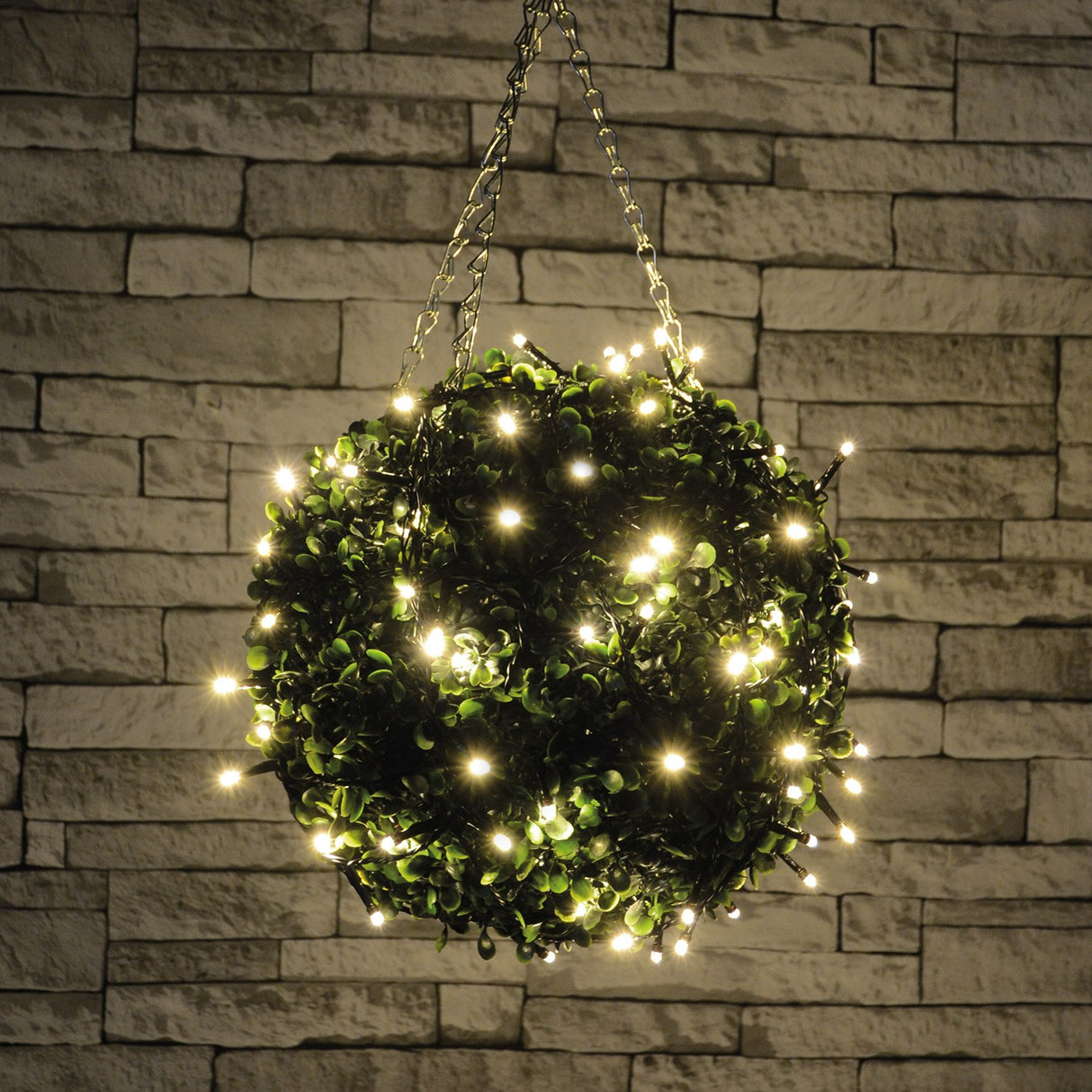 LYYT topiary ball light with 120 warm white LEDs wrapped around a green foliage sphere, hung by chains against a stone brick wall.