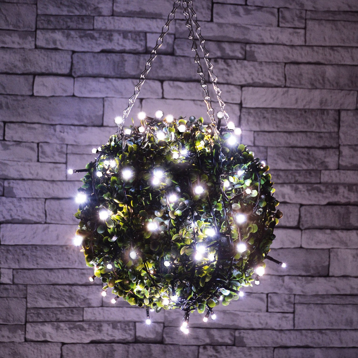 LYYT LED Battery Topiary Ball Light with 120 cool white LEDs wrapped around a green foliage sphere, hung by chain against a grey brick wall.