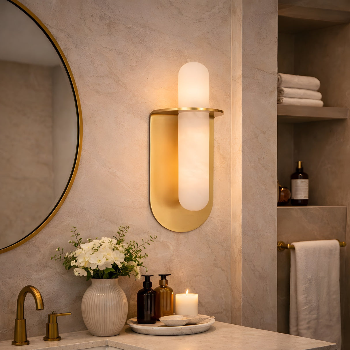 Modern bathroom with a round mirror, gold faucet, Luneth LED Small Alabaster Wall Light in Brushed Brass & Soft White, white flower vases, soap dispensers, a lit candle, and neatly stacked towels. Elegant and neutral-toned decor.