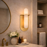 Modern bathroom with a round mirror, gold faucet, Luneth LED Small Alabaster Wall Light in Brushed Brass & Soft White, white flower vases, soap dispensers, a lit candle, and neatly stacked towels. Elegant and neutral-toned decor.
