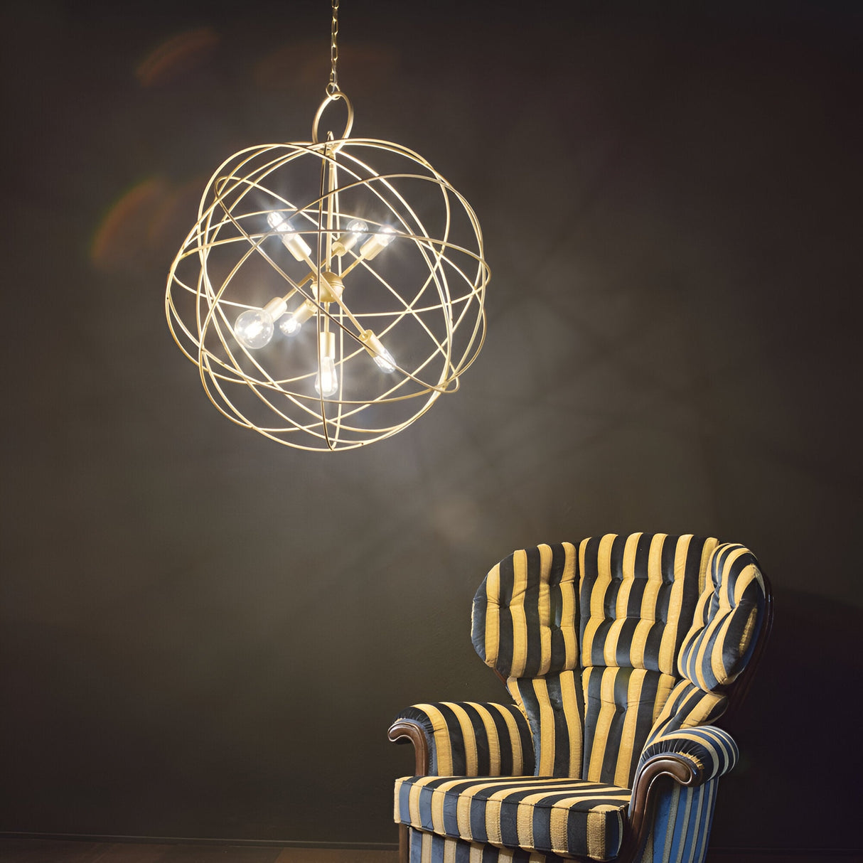 A Zen 7 Light Suspension in Gold with an ornate geometric design and exposed bulbs hangs from the ceiling, casting light over a cozy corner featuring a striped armchair against a dark wall.