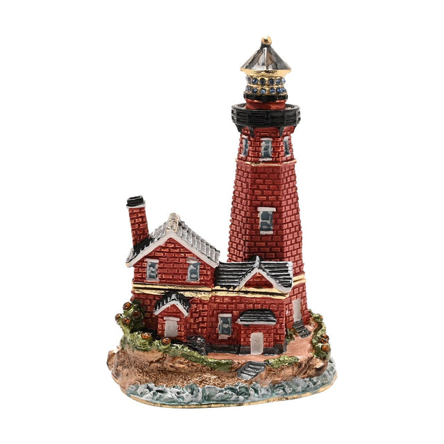 Painted metal trinket box shaped as a red brick lighthouse with a keeper's cottage, gold banding, grey roofs, and a black lantern gallery set with blue stones, on a plain white background.