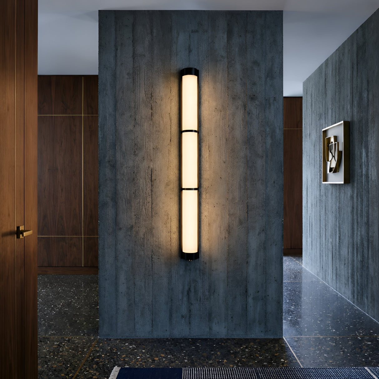 A modern hallway features the Arelis LED Linear Alabaster Wall Light in soft white & black, mounted on a textured concrete wall, with wood-paneled walls, terrazzo flooring, and a framed abstract artwork.