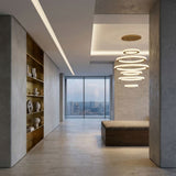 A modern, minimalist hallway with beige stone floors, built-in wooden shelves displaying white ceramics, a cushioned bench, the Celine LED 6 Tier Suspension Light in gold, and large windows overlooking a city skyline.