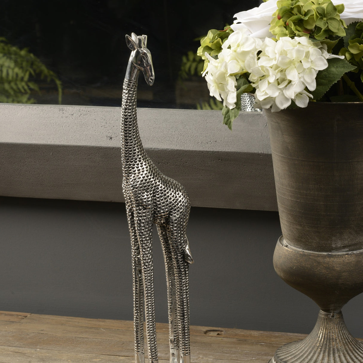 Libra Interiors Textured Electroplated Giraffe Sculpture in silver resin, displayed on a wooden surface beside a bronze urn of white hydrangeas.