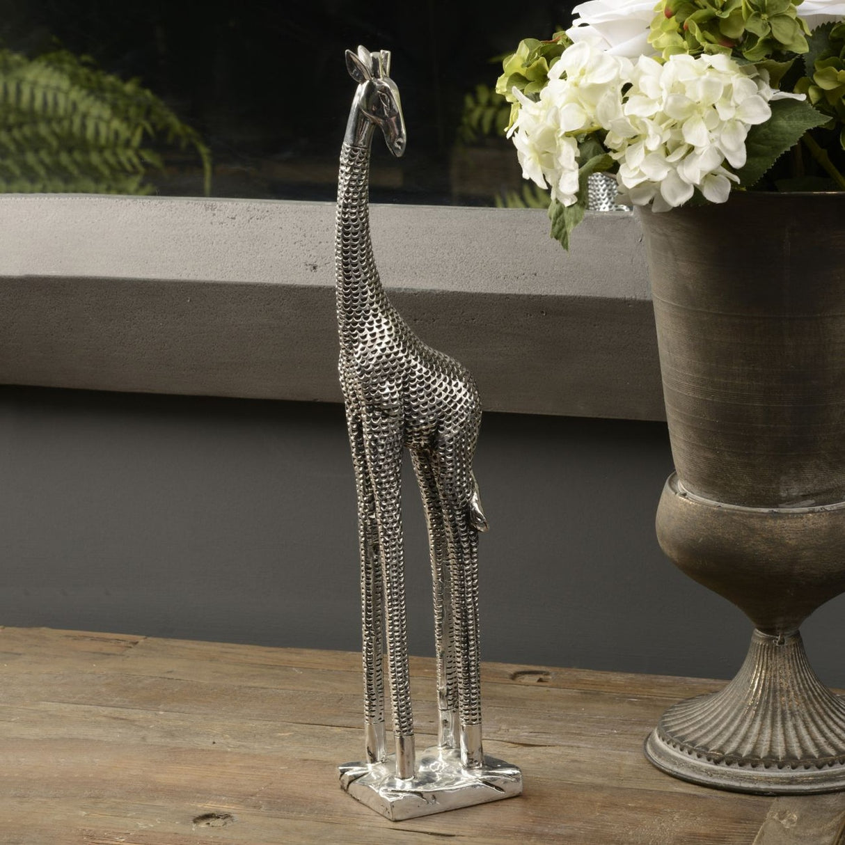 Libra Interiors Textured Electroplated Giraffe Sculpture in silver resin on a wooden surface beside a metal urn of white hydrangeas.