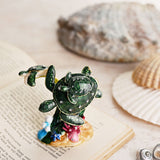 Coral Turtle Metal Trinket Box with Hinged Shell Lid in glossy green, shown with sea shells and an open book, featuring a large turtle carrying a baby turtle and a seaweed frond with a smaller turtle.