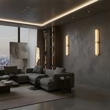 A modern living room with gray walls, city views through large windows, two Eliora LED Alabaster Wall Lights in brushed brass and soft white, a gray sectional sofa, coffee table, decor shelves, and soft lighting for a cozy feel.