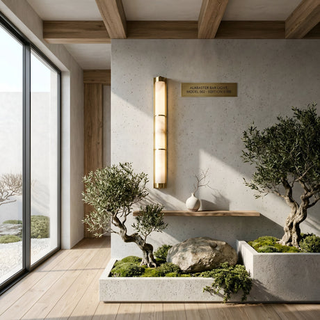A minimalist interior with a large window features a compact zen garden with bonsai, moss, and stones in concrete planters. The Arelis LED Vertical Alabaster Wall Light in Soft White & Brushed Brass and decorative vases adorn the textured wall.