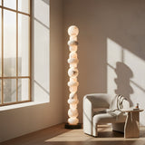 The Stratalux LED Stacked Column Alabaster Floor Lamp in matte black and soft white stands by a cozy white armchair with a blanket, next to a small wooden table in a sunlit minimalist room with large windows and light walls.