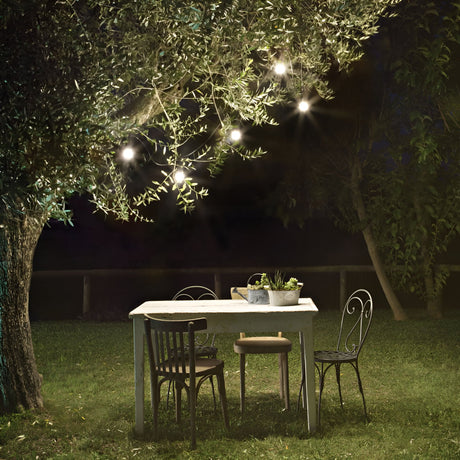 In a dimly lit outdoor setting, a rustic wooden table surrounded by mismatched chairs rests on the grass beneath a tree. From the branches above, Velox Outdoor String Lights in black, stretching 4 meters and featuring energy-efficient LED bulbs, emit a gentle glow. A potted plant placed on the table enhances the cozy and serene ambiance.