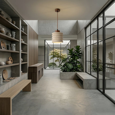 A modern minimalist space with concrete floors, walnut brown built-in shelves, a wooden bench, large windows framing a lush indoor garden, and the Rivano 1 Light Ribbed Drum Travertine Single Pendant Light in Walnut Brown & Light Beige hanging overhead.