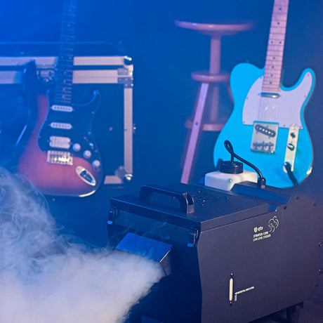 QTX Stratus 1200 low level fog machine emitting dense ground-hugging white fog on a stage set with electric guitars, flight case and stool under blue light.