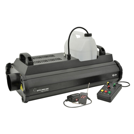 The QTX QTFX-2000 Mkii High Power Fog Machine - 2000W is a large black unit featuring a detachable jug on top, intelligent temperature regulation, and DMX control. It comes with a remote and two attached control boxes equipped with buttons and dials.