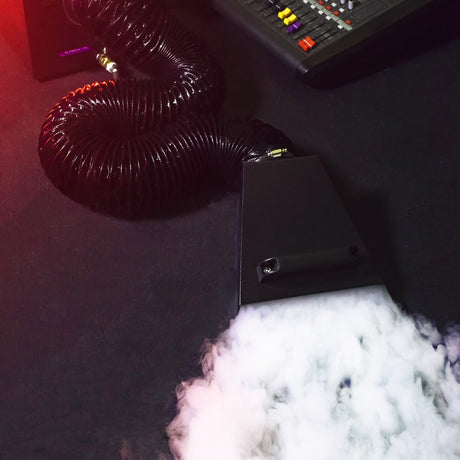 QTX UMBRA-1200 low fog machine by AVSL emitting dense ground-hugging mist through its flexible duct beside a lighting control desk.