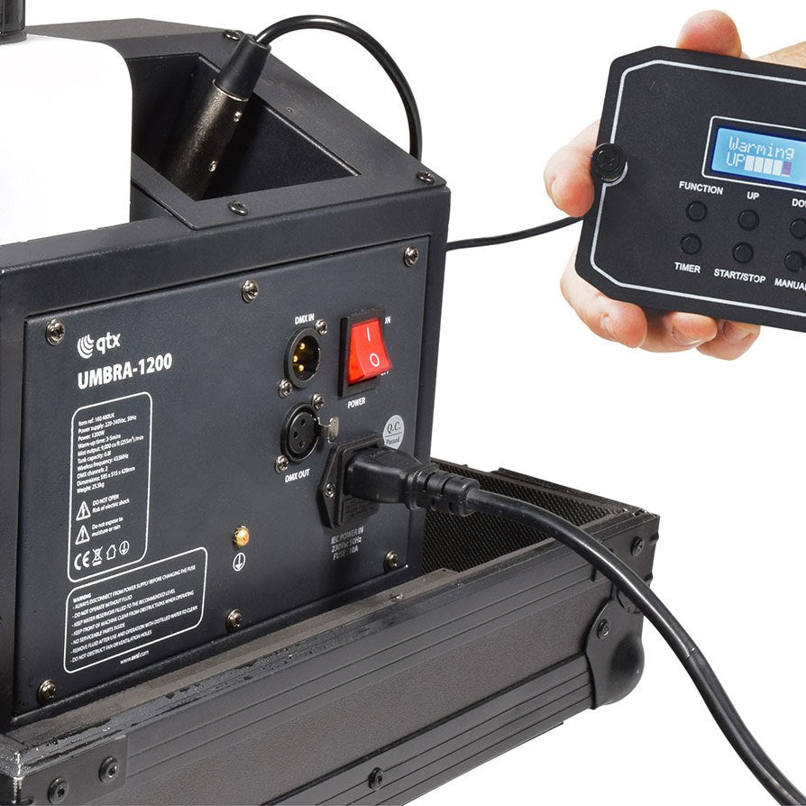 QTX UMBRA-1200 low mist generator rear panel showing DMX in/out XLR ports, red power switch, IEC inlet and detachable digital remote displaying Warming Up.