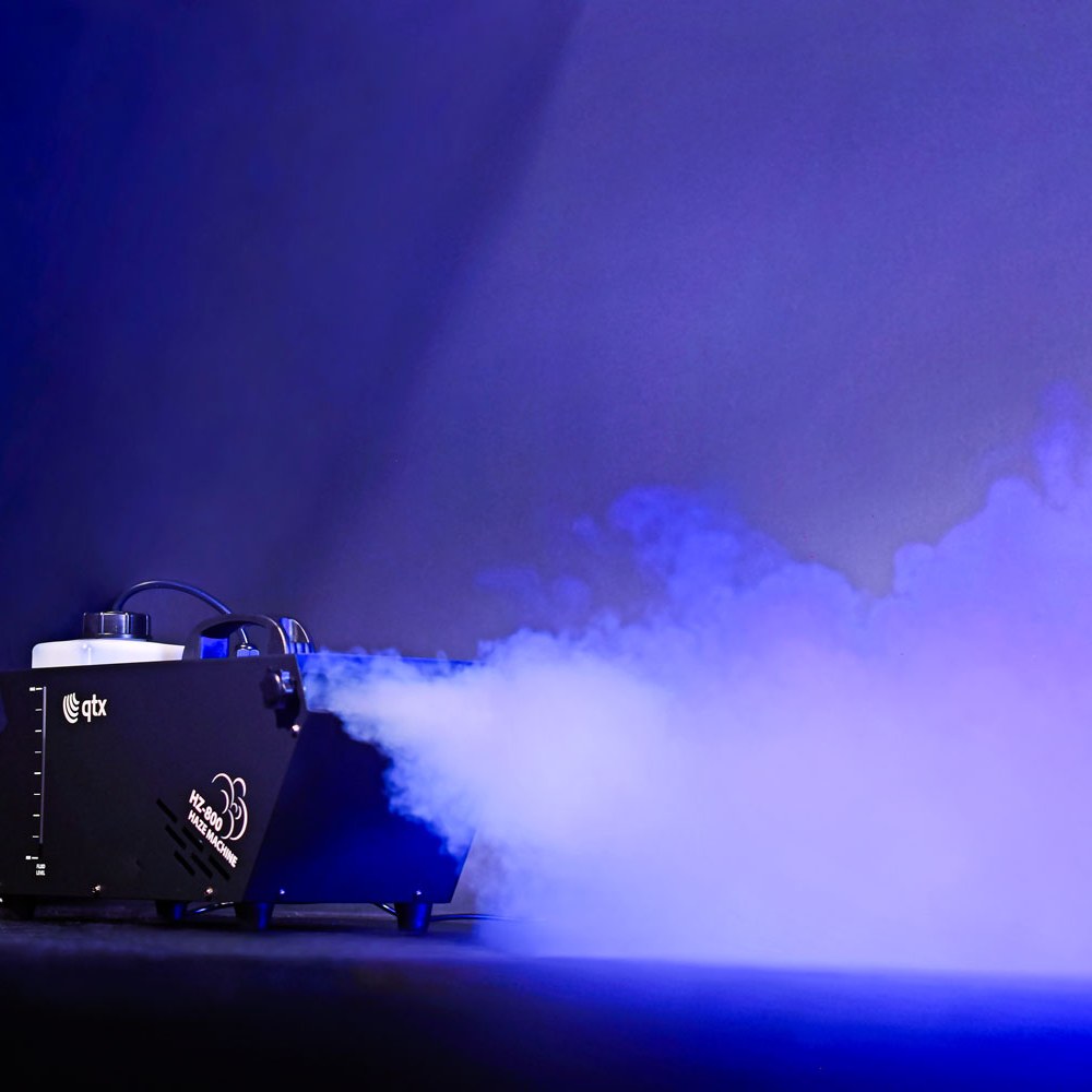 QTX HZ-800 800W haze machine in black metal casing with fluid tank, emitting dense haze under blue stage lighting against a dark background.