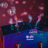 QTX Tetra Bubble 4-wheel bubble machine in action, streams of bubbles rising from the black casing under red and blue stage lighting.