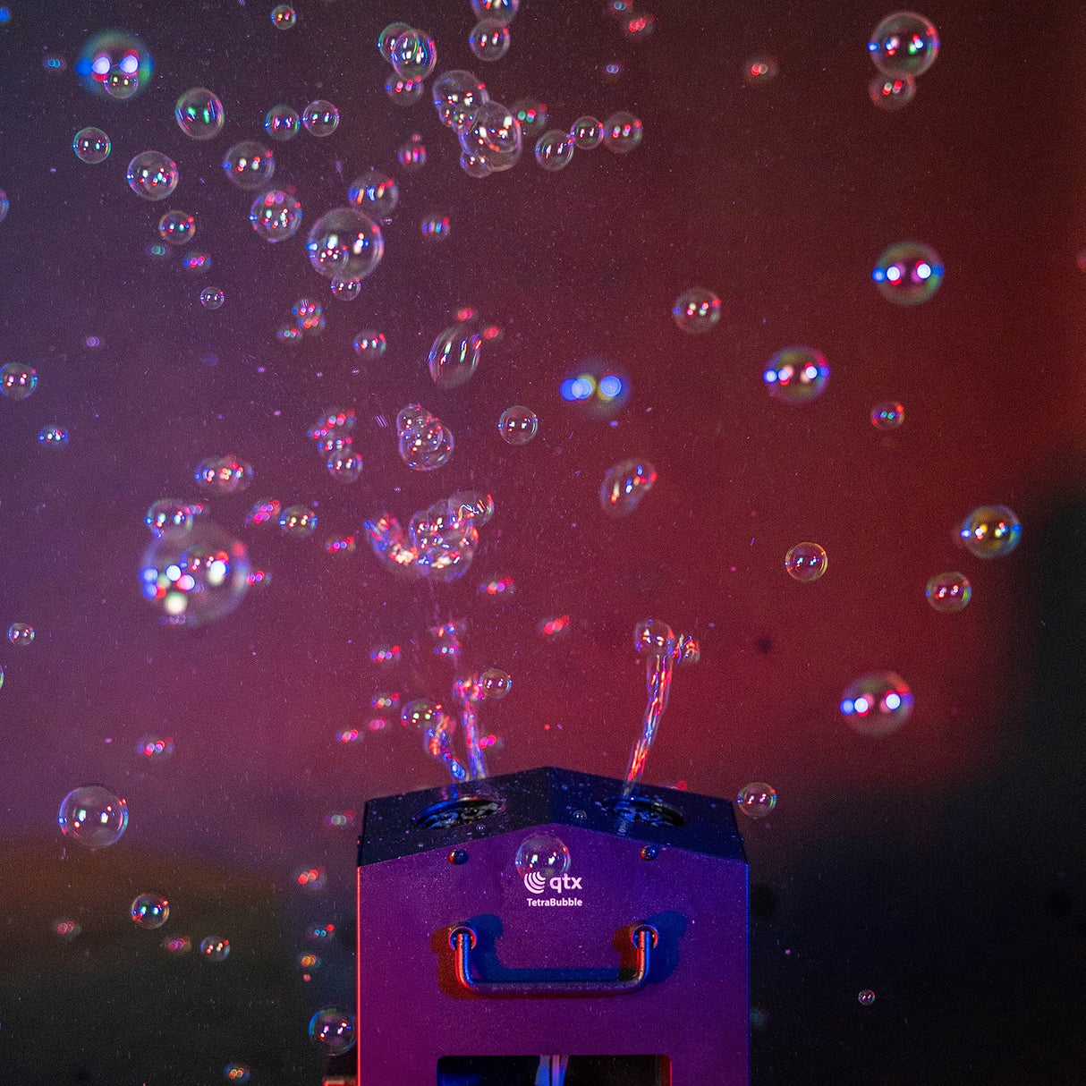 QTX Tetra Bubble machine in use, streaming bubbles upward from its four wheels against a dark red and purple lit background.