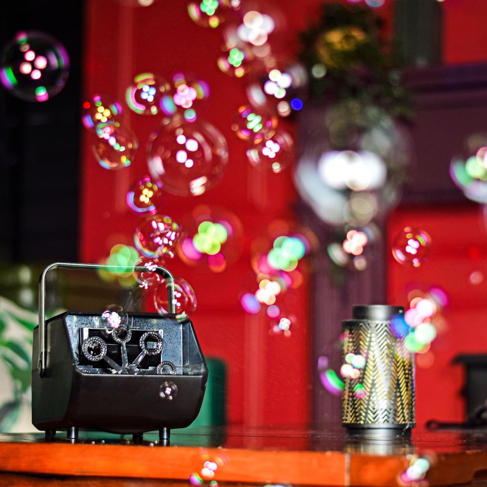 AVSL QTX Mini Bubble USB compact black bubble machine with carry handle and dual wheels on a wooden surface, releasing bubbles beside a metal lantern.