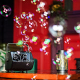 AVSL QTX Mini Bubble USB compact black bubble machine with carry handle and dual wheels on a wooden surface, releasing bubbles beside a metal lantern.