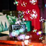 QTX Mini Bubble USB compact bubble machine with carry handle on a wooden table, releasing colourful bubbles against a red and green backdrop.