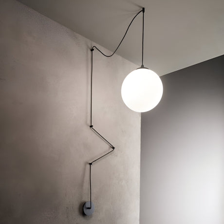 The Sense 1 Light Wall/Pendant in black is suspended from the ceiling, its cord weaving along a textured beige wall. This modern spherical design perfectly suits contemporary interiors, casting an ambient glow that stands out against the smooth, gray surface of the nearby wall.