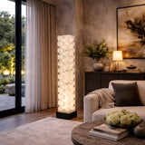 A modern living room features the Hexaryn LED Interlocking Alabaster Floor Lamp in Matte Black & Soft White glowing beside a white sofa, with a round coffee table, sideboard decor, and ambient light near large windows with sheer curtains.