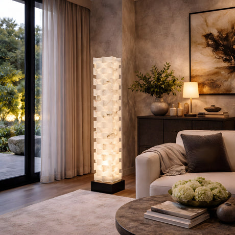 A modern living room features the Hexaryn LED Interlocking Alabaster Floor Lamp in Matte Black & Soft White glowing beside a white sofa, with a round coffee table, sideboard decor, and ambient light near large windows with sheer curtains.