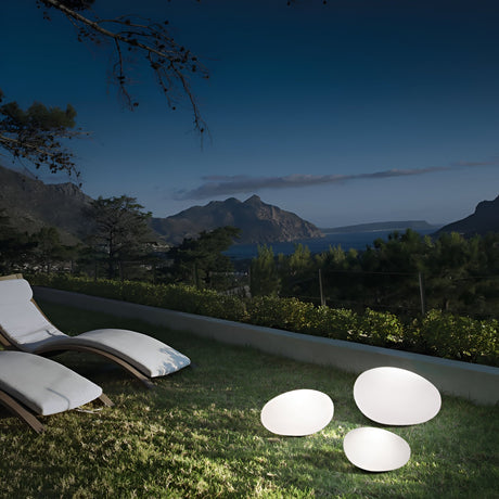 A serene night view showcases lounge chairs on a grassy lawn, overlooking mountains and a body of water. In the foreground, three Zest Outdoor Pedestal Lights in white, each standing 25cm tall with opal plastic diffusers, cast a soft glow across the grass.