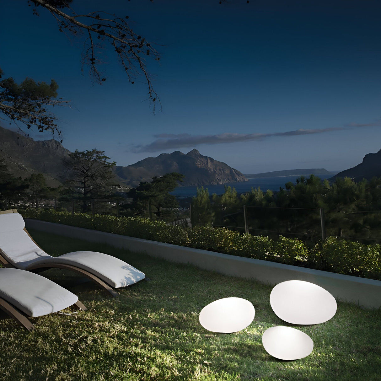 A serene nighttime landscape showcases a grassy yard illuminated by three glowing, pebble-shaped lights. The scene is enhanced with elegant Zest Outdoor Pedestal Lights in white, standing 41cm tall. Two lounge chairs are positioned nearby, offering a stunning view of distant mountains and the sea under a clear, starry sky.