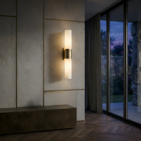 The Velaris 2 Light Double Alabaster Wall Light in soft white and brass is mounted on a textured beige wall next to a low cabinet. Large windows showcase an evening view of trees and stone structures outside.