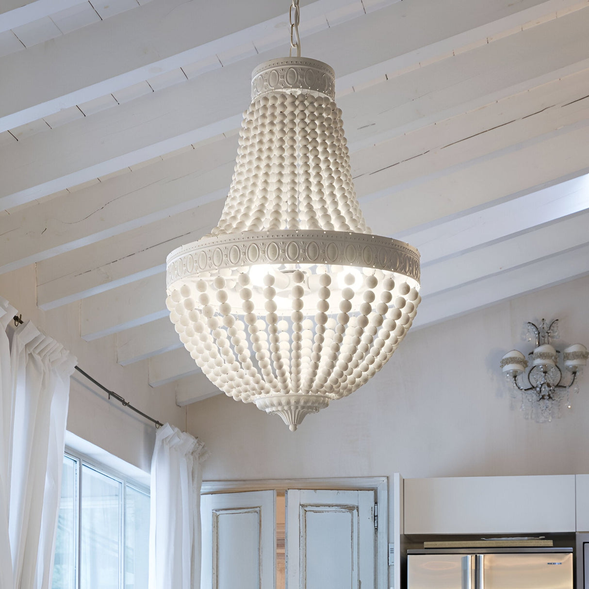 A large, elegant Sync Wood 6 Light Chandelier - White hangs from the ceiling in a bright room, featuring natural wooden beads. The ceiling boasts white wooden beams, and sheer white curtains by the window enhance the airy atmosphere, perfectly capturing modern farmhouse décor.