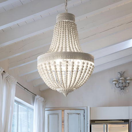 A large, elegant Sync Wood 6 Light Chandelier - White hangs from the ceiling in a bright room, featuring natural wooden beads. The ceiling boasts white wooden beams, and sheer white curtains by the window enhance the airy atmosphere, perfectly capturing modern farmhouse décor.