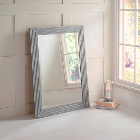 The Pearl Rectangle Wall Mirror, with its textured grey and silver frame, leans against a white paneled wall beside a window. A candle in a glass holder is placed on a stack of books on the floor next to the mirror, adding an elegant touch to the home decor.