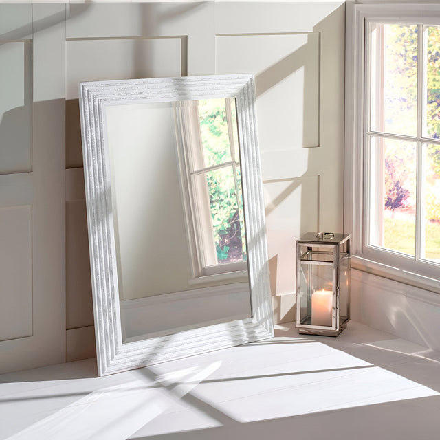 A Pearl Rectangle Wall Mirror - White/Silver leans against a white paneled wall, accompanied by a glass lantern holding a lit candle. Sunlight filters through a nearby window, creating shadows on the wall and floor and enhancing the modern elegance of the scene.