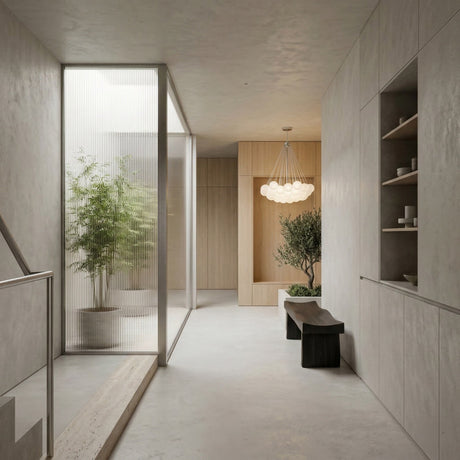 Minimalist hallway with light walls, built-in shelves, a black bench, and an indoor plant. A glass partition separates another area featuring a large potted plant and the Orb 37 Light Cluster Suspension - Silver with frosted glass spheres overhead.