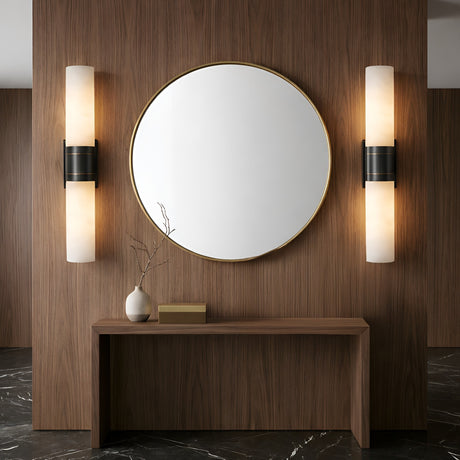 A modern entryway features a round gold-framed mirror on a wooden wall, flanked by Velaris 2 Light Double Alabaster Wall Lights in Soft White & Black. Below is a narrow wooden console with a white vase holding bare branches and a rectangular box.